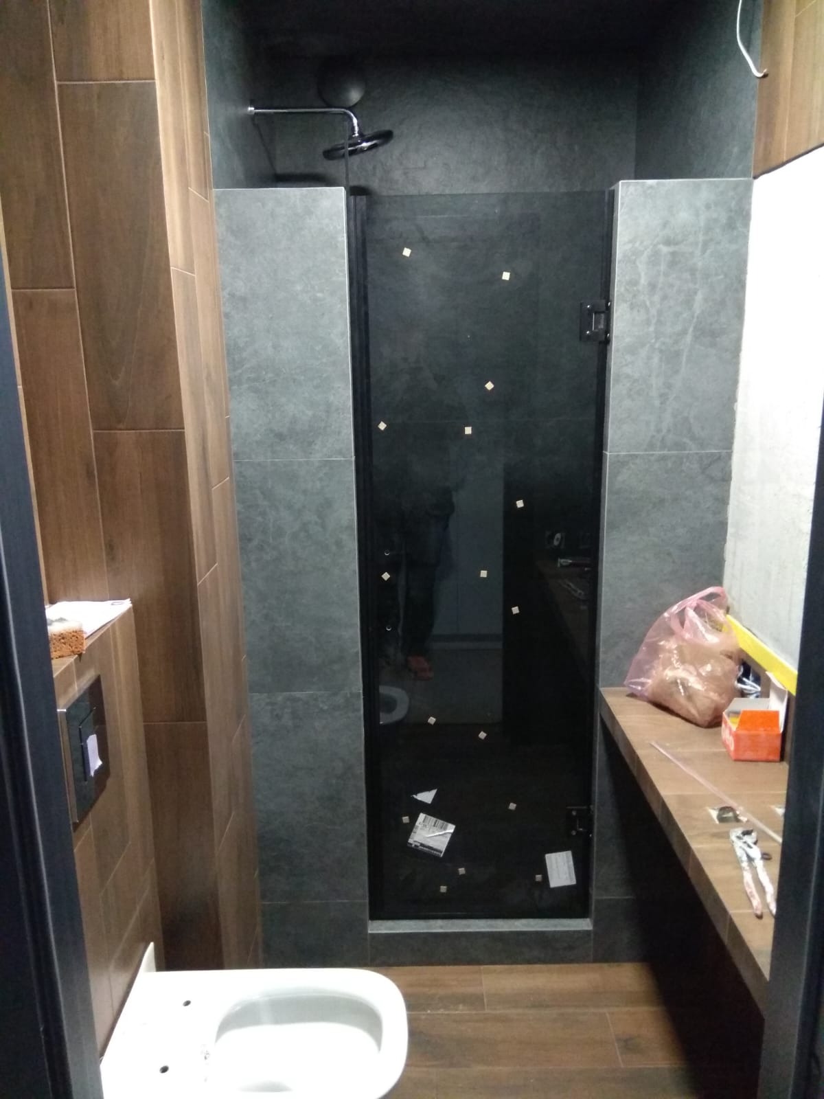 Nowoczesna kabina prysznicowa z czarnymi drzwiami i geometrycznym wzorem, szare płytki, drewnopodobne panele podłogowe, fragment toalety i blatu z narzędziami.