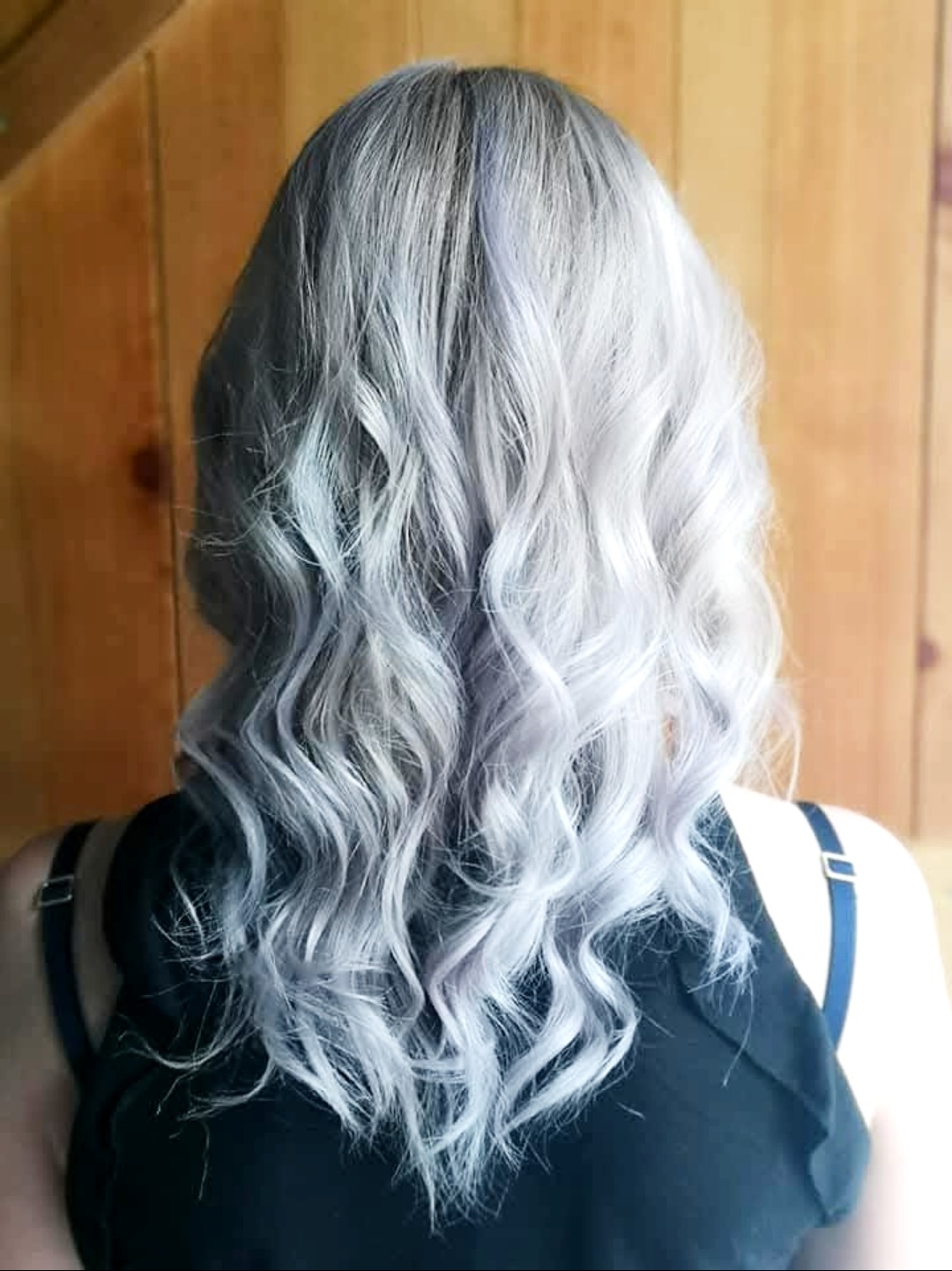 Tył głowy kobiety z pofalowanymi włosami w odcieniach szarości i bieli, z delikatnym fioletowym połyskiem, na tle drewnianej ściany.