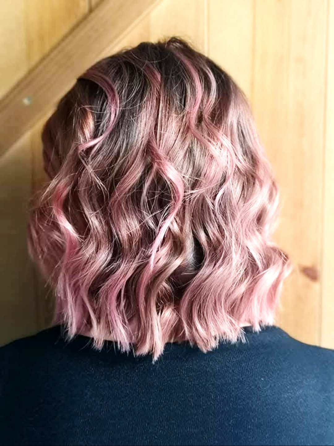 Falowane, krótkie włosy z różowymi pasemkami, widok z tyłu, ciemny sweter, drewniane drzwi w tle. Świeża koloryzacja z efektem ombre.