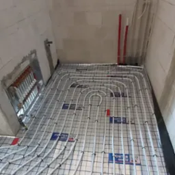 Białe rury ogrzewania podłogowego ułożone na srebrnej folii z nadrukiem, w pomieszczeniu z białymi ścianami z bloczków. Widoczny rozdzielacz instalacji.