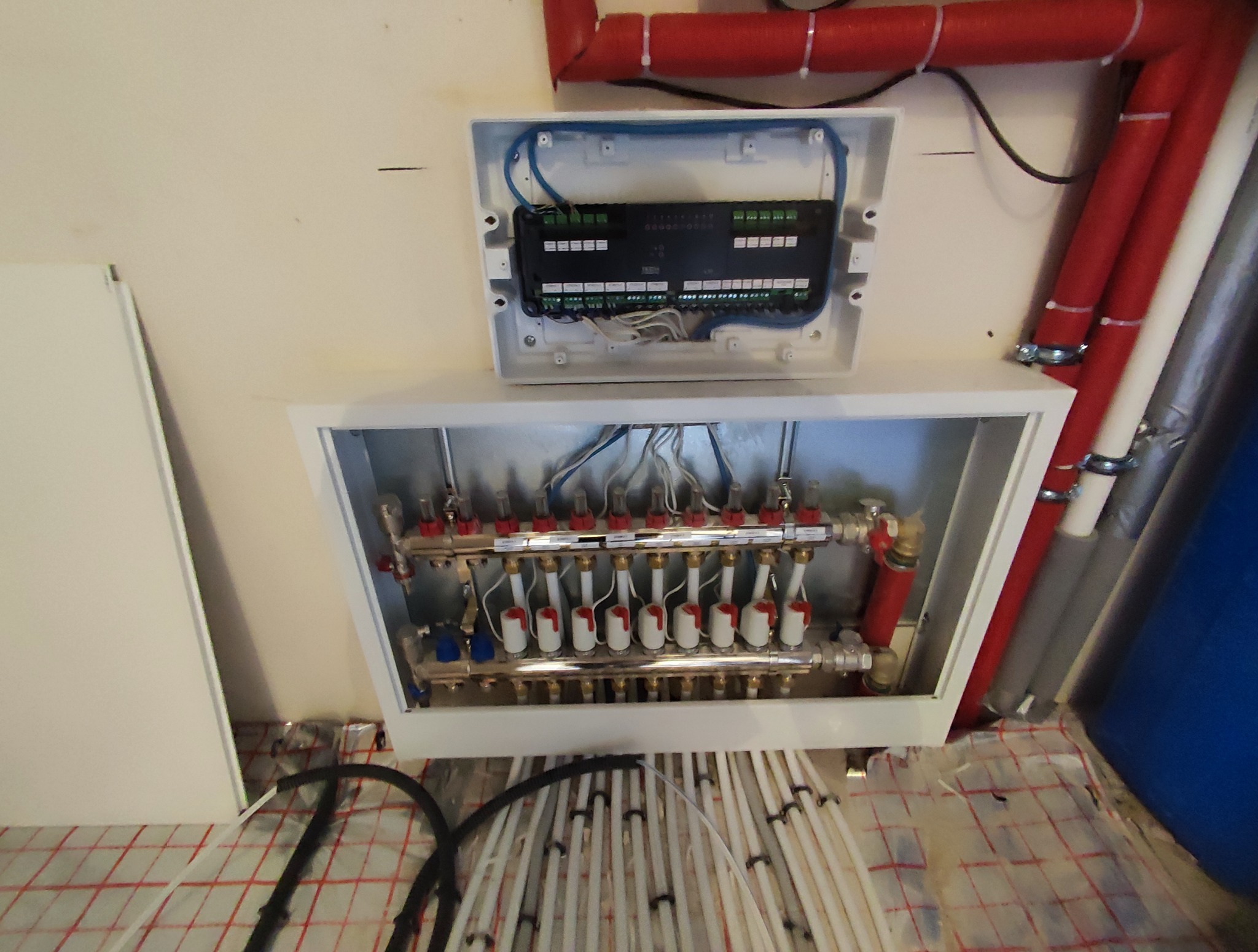 Rozdzielacz ogrzewania podłogowego w szafce, z widocznymi rurami i elementami sterującymi. Instalacja w trakcie montażu na siatce grzewczej.