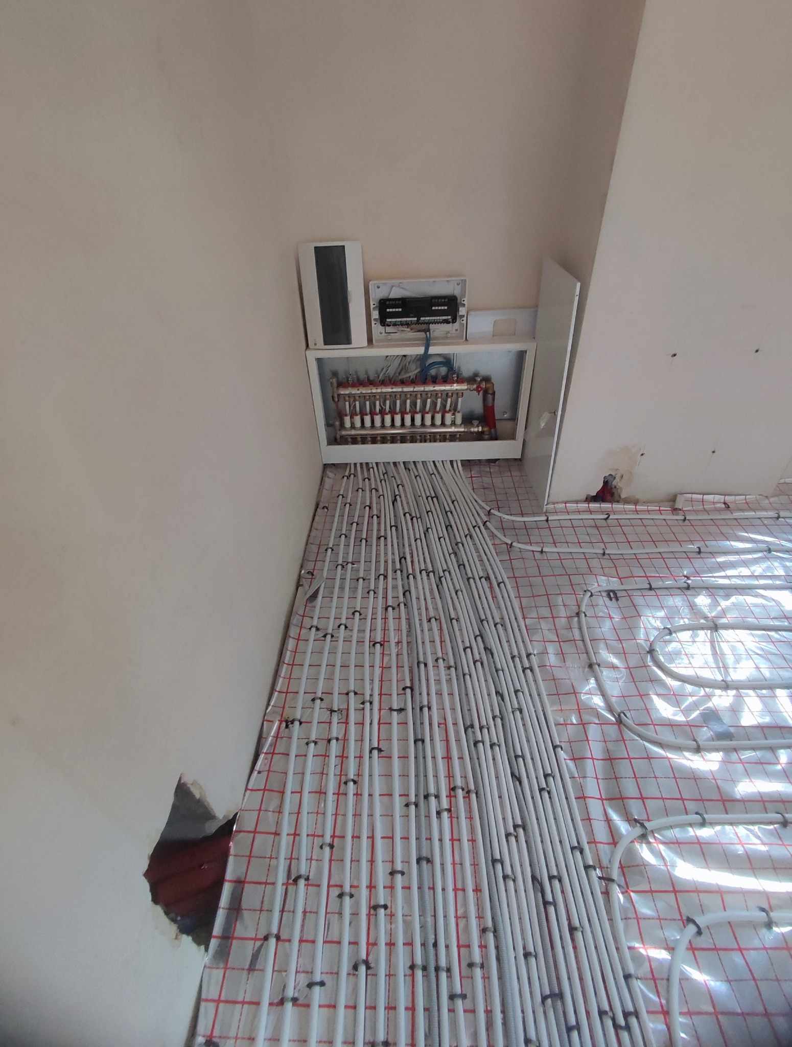 Białe rury ogrzewania podłogowego ułożone na folii z czerwoną siatką, widoczny rozdzielacz w szafce na ścianie. Instalacja w trakcie montażu.