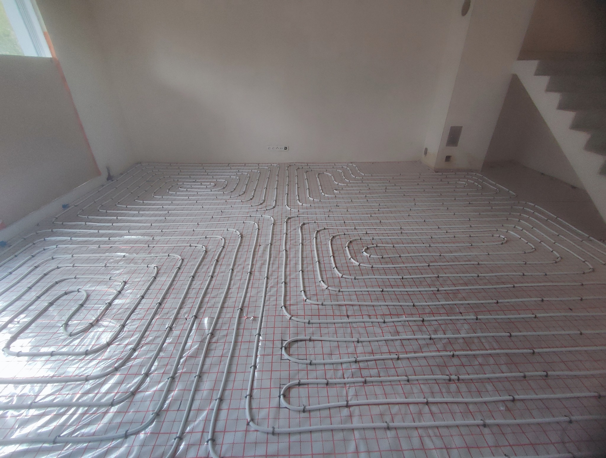 Białe rury ogrzewania podłogowego ułożone spiralnie na czerwonej siatce w jasnym, surowym pomieszczeniu z widocznymi schodami w tle.