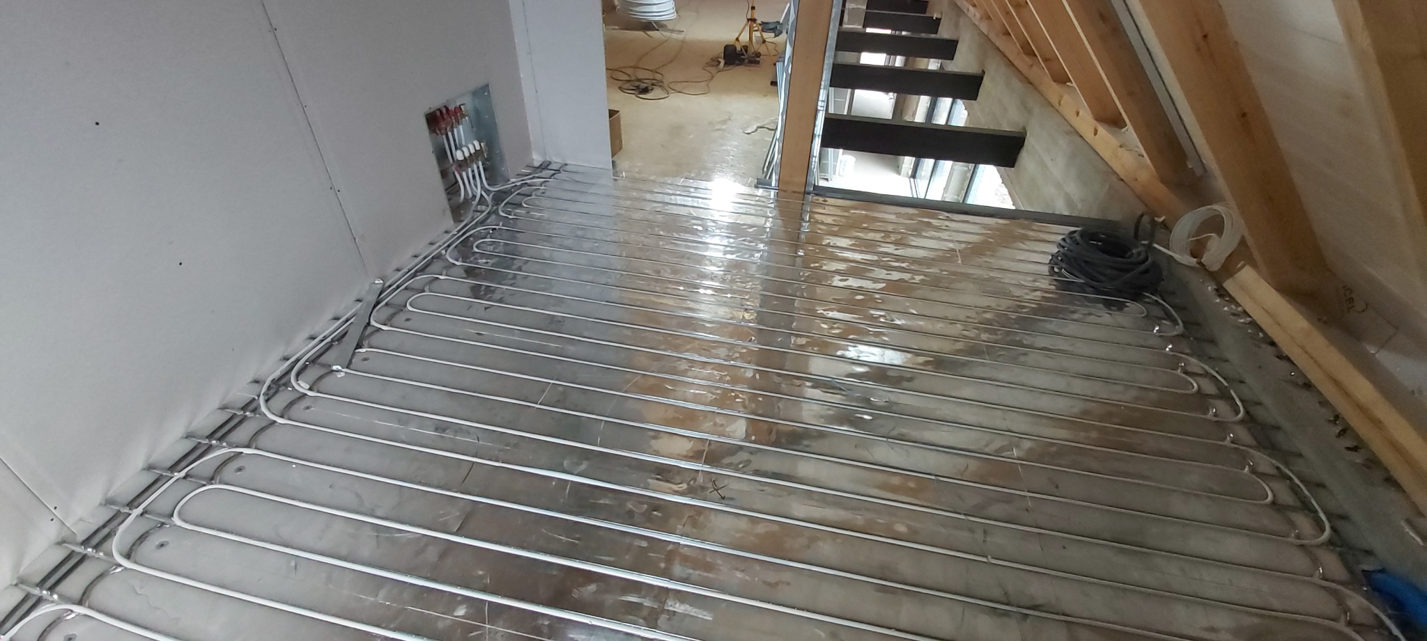 Ułożone rury ogrzewania podłogowego na folii izolacyjnej w surowym wnętrzu poddasza z widocznymi schodami i instalacją przy ścianie.