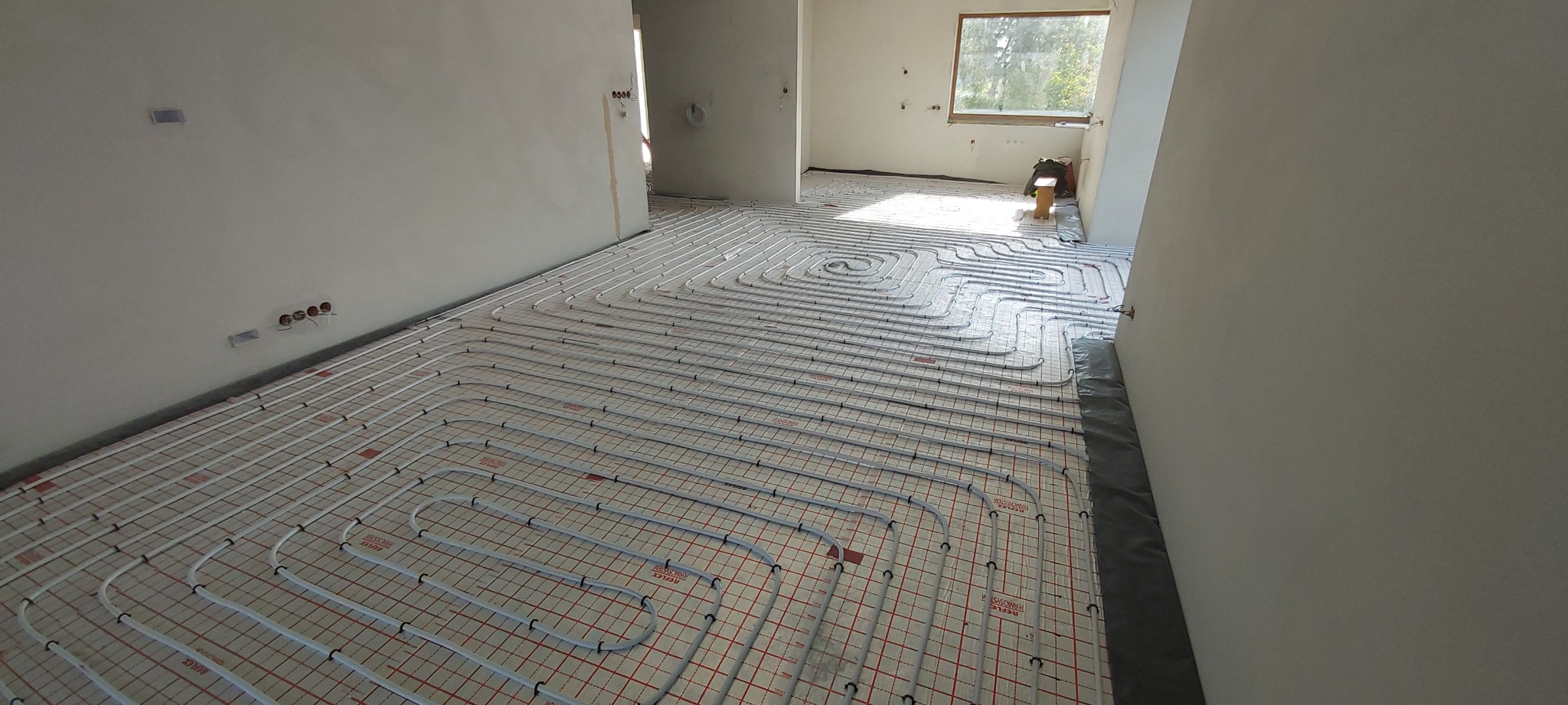 Białe rury ogrzewania podłogowego ułożone na czerwonej macie w surowym wnętrzu z oknem. Widoczne wystające kable elektryczne.