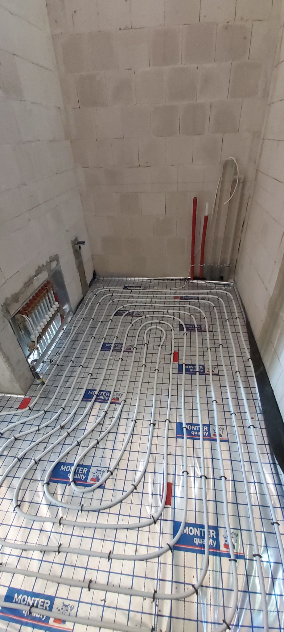 Białe rury ogrzewania podłogowego ułożone na srebrnej folii z nadrukiem, w pomieszczeniu z białymi ścianami z bloczków. Widoczny rozdzielacz instalacji.