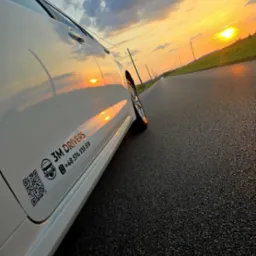 Biały samochód z logo 3M Drivers na tle zachodzącego słońca i drogi. Widoczny numer telefonu i kod QR. Perspektywa ukośna z bliska, akcent na detale pojazdu.
