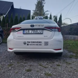 Biała Toyota Corolla z oznaczeniami Taxi 3M Drivers, widoczna z tyłu na żwirowym podłożu. Tablica rejestracyjna GA 786LU. W tle dom i drzewa.