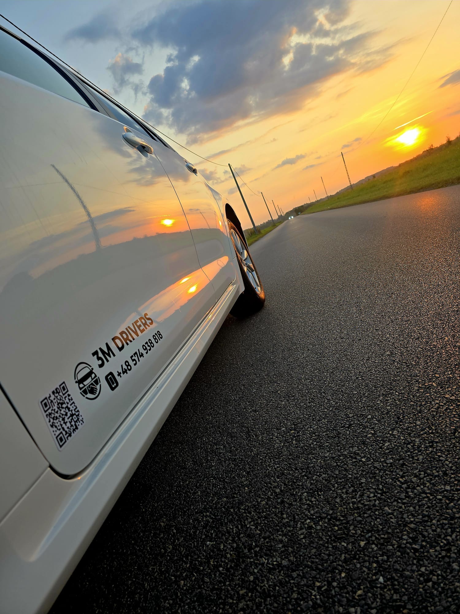 Biały samochód z logo 3M Drivers na tle zachodzącego słońca i drogi. Widoczny numer telefonu i kod QR. Perspektywa ukośna z bliska, akcent na detale pojazdu.