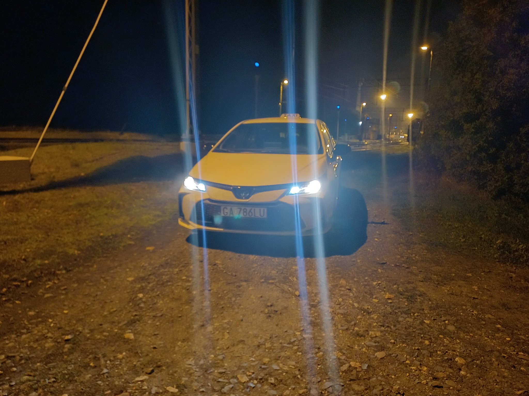 Żółta taksówka z włączonymi światłami na żwirowej drodze nocą, widoczne lampy uliczne i drzewa w tle. Tablica rejestracyjna pojazdu widoczna.