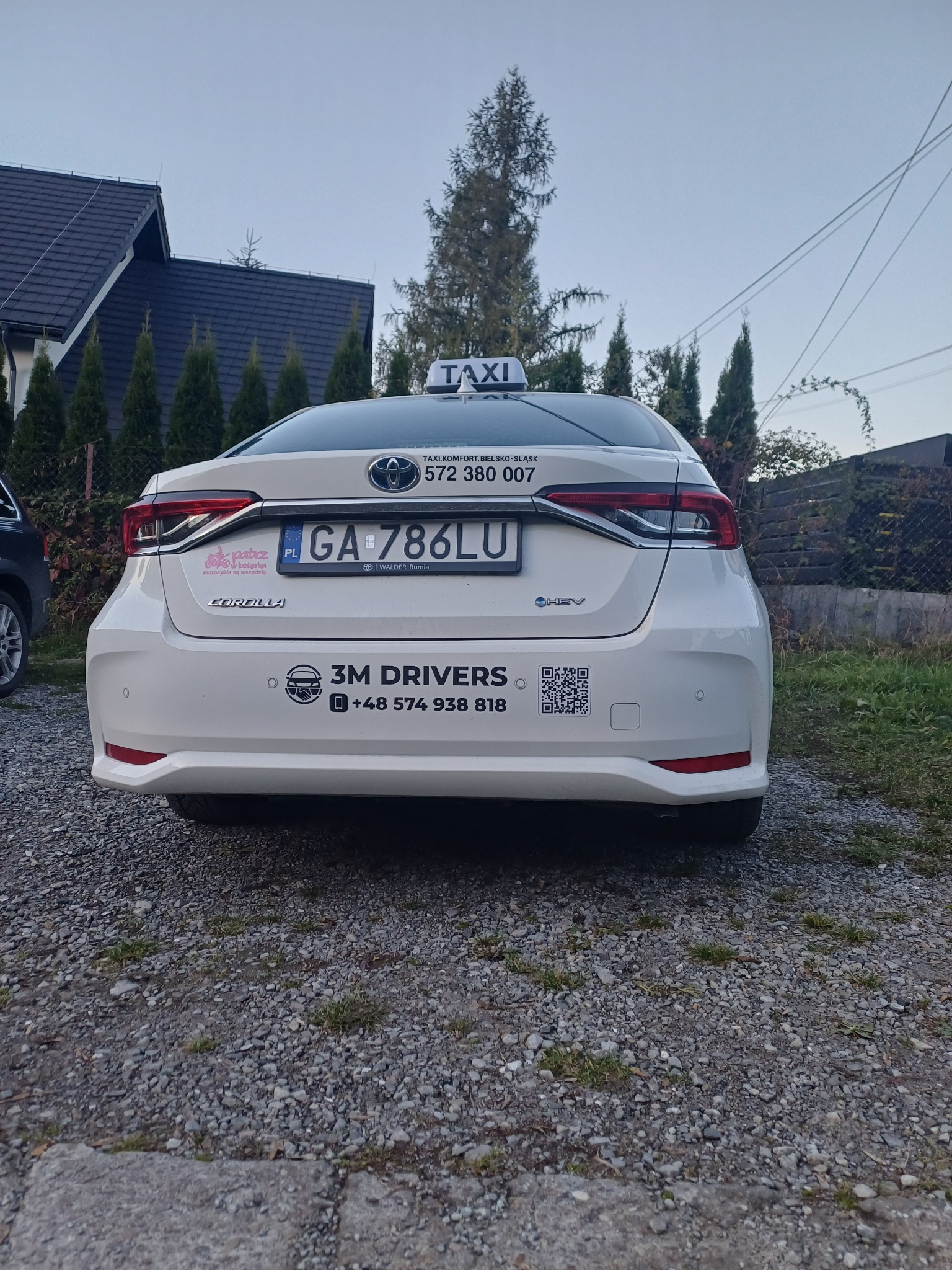 Biała Toyota Corolla z oznaczeniami Taxi 3M Drivers, widoczna z tyłu na żwirowym podłożu. Tablica rejestracyjna GA 786LU. W tle dom i drzewa.