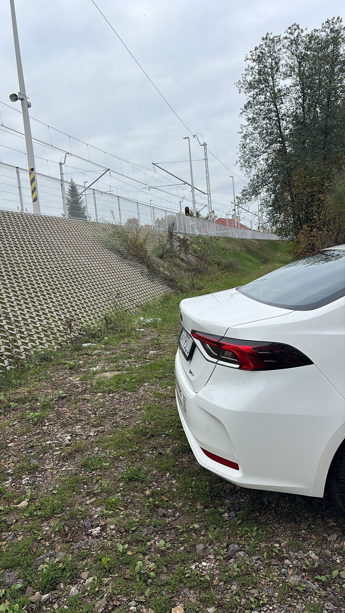 Biały samochód osobowy zaparkowany obok torów kolejowych, widok z tyłu, pochmurne niebo w tle. Widoczny fragment tablicy rejestracyjnej.