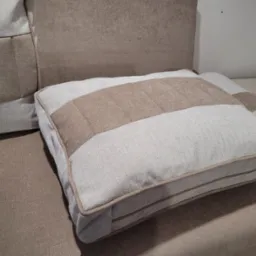 Beżowa poduszka dekoracyjna z pasami na tapicerowanym siedzisku, w tle fragment sofy i maszyna do szycia. Widoczne detale szwów i faktura tkaniny.