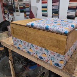 BMT TAPICER Patryk Kowalczuk - Drewniana skrzynia z tapicerowanym siedziskiem w dinozaury, stoi na warsztacie. W tle wzorniki tkanin. Widoczny fragment narzędzia.