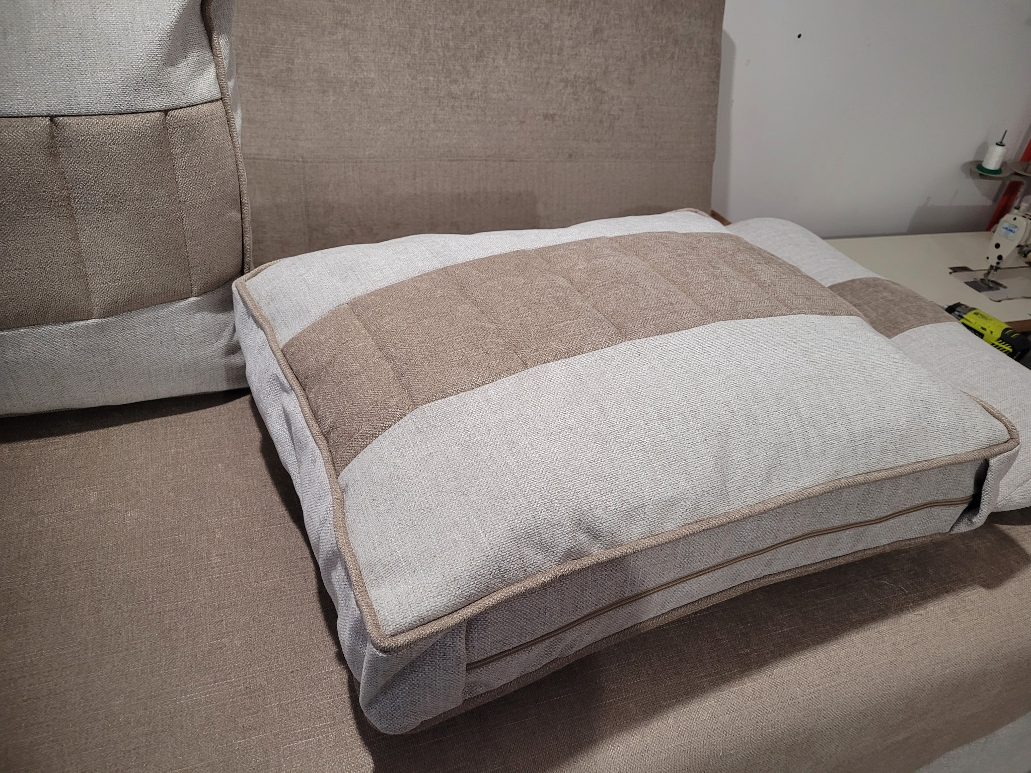 Beżowa poduszka dekoracyjna z pasami na tapicerowanym siedzisku, w tle fragment sofy i maszyna do szycia. Widoczne detale szwów i faktura tkaniny.