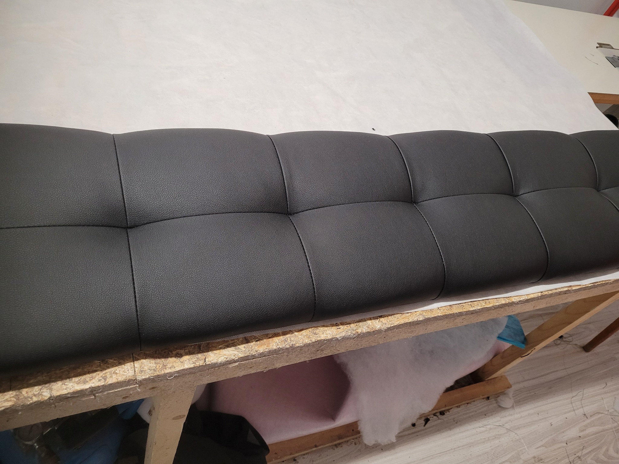 Czarny, pikowany panel tapicerowany na drewnianym stelażu, widoczne wypełnienie z waty. Detal z warsztatu tapicerskiego, bliski kadr.