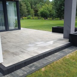 Szymon Niżnikiewicz - Nowoczesny taras z jasnoszarej kostki brukowej, obrzeża z ciemnoszarego granitu. Widok na zadbany trawnik i zieleń ogrodu. Minimalistyczny design.