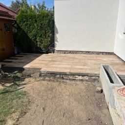 Szymon Niżnikiewicz - Układanie jasnej kostki na tarasie przy budynku. Widoczna ubijarka, przygotowany teren i fragment betonowej konstrukcji. Prace brukarskie w toku, słoneczny dzień.