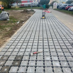 Szymon Niżnikiewicz - Szara kostka brukowa ECO-BRUK ułożona na ścieżce, z ubijarką i gumowym młotkiem w tle, w otoczeniu zieleni i budynków miejskich. Widok z góry.