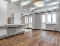 Jasne, przestronne wnętrze po remoncie: parkiet w ciepłym odcieniu brązu, białe ściany, grzejnik pod oknem i nowoczesne oświetlenie sufitowe. Widok na aneks kuchenny.