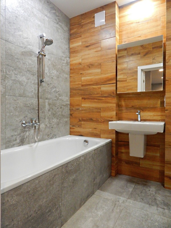 Wykończona łazienka z wanną obudowaną szarą płytką, drewnopodobnymi kaflami na ścianie, białą umywalką i lustrem. Widoczny fragment szarej podłogi.