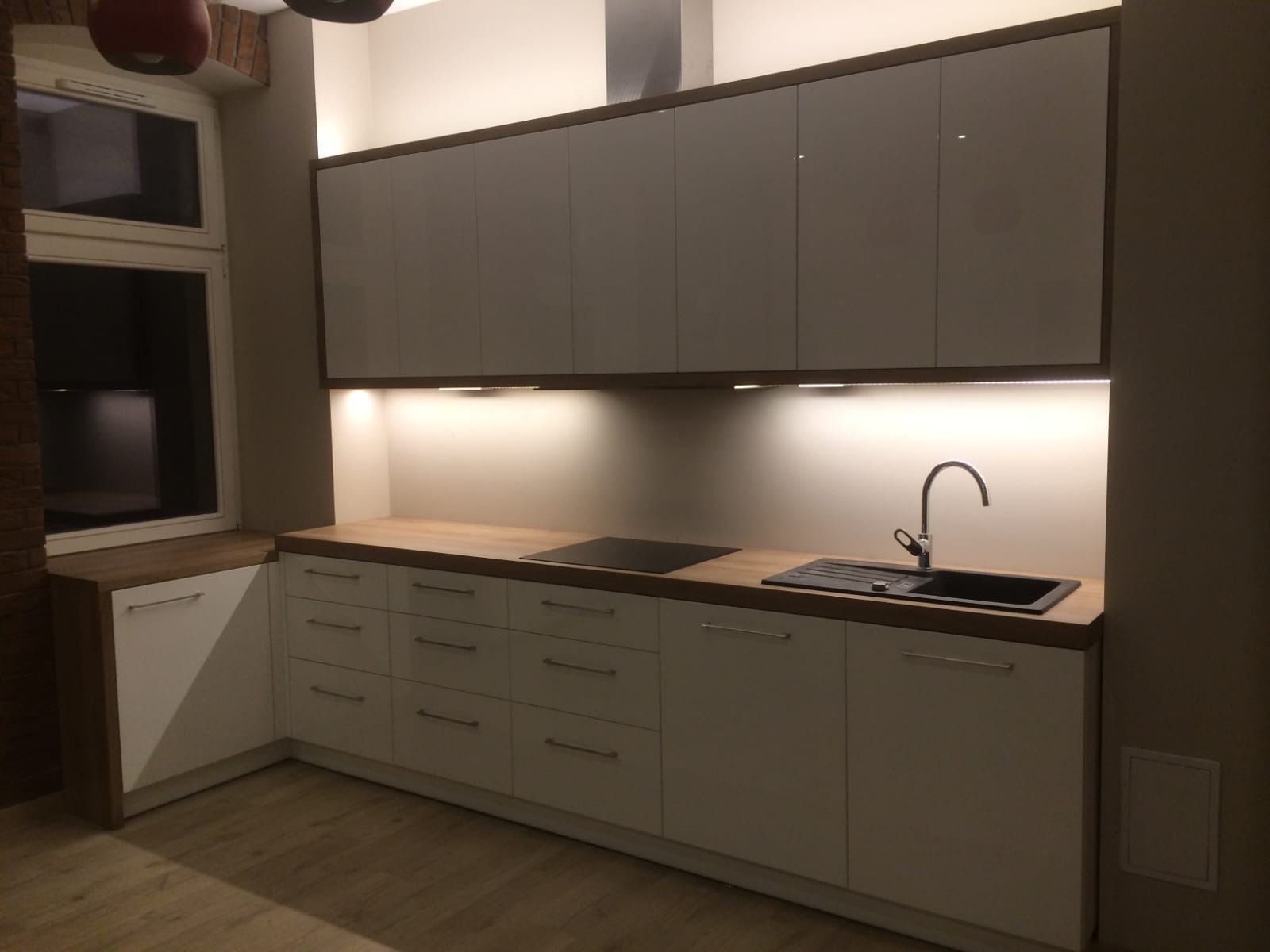 Nowoczesna kuchnia z szarymi szafkami górnymi z ciemnym obramowaniem i białymi dolnymi, drewniany blat, czarny zlew i płyta, oświetlenie LED podszafkowe.