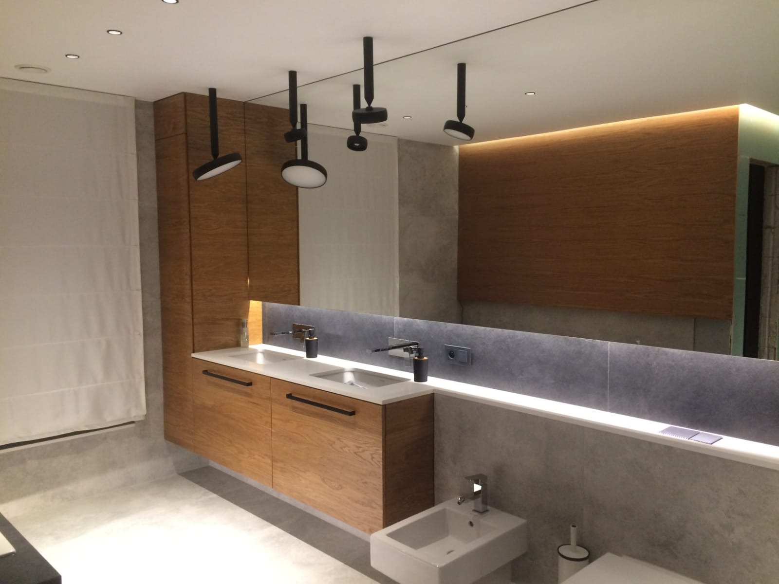 Nowoczesna łazienka z drewnianą szafką pod umywalką, podświetlanym lustrem i czarnymi lampami sufitowymi. Minimalistyczny design z elementami drewna.