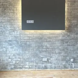 Ściana z szarej cegły z podświetlanym panelem i gniazdkami, widoczne drewniane belki stropowe. Parkiet w trakcie renowacji. Kabel na podłodze.