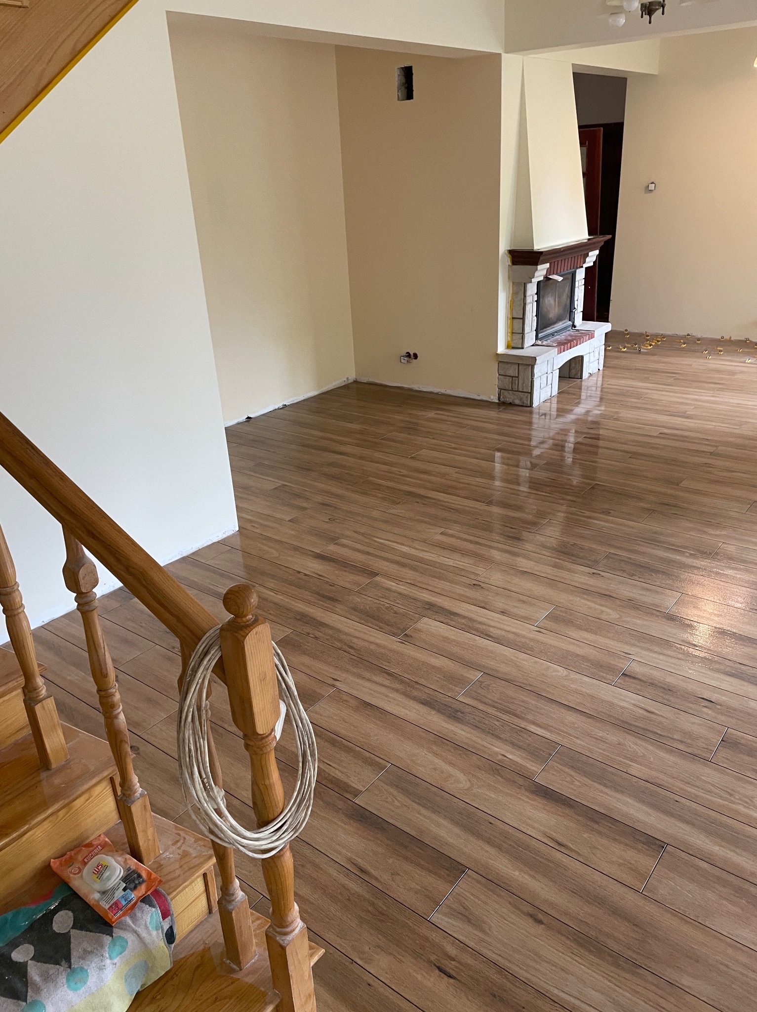 Ukończona podłoga z płytek imitujących drewno w salonie z kominkiem. Widoczne schody i akcesoria glazurnicze na pierwszym planie. Jasne ściany i dobre oświetlenie.