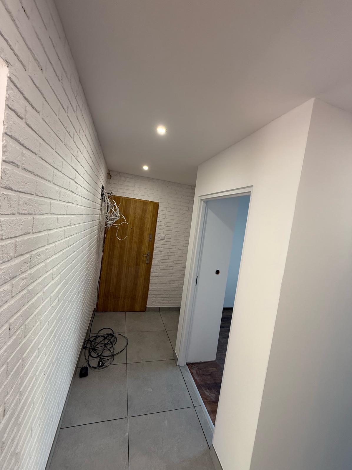 Korytarz po remoncie: biała cegła na ścianie, szara podłoga, drewniane drzwi i białe ściany z wejściem do pokoju. Widoczne kable elektryczne.