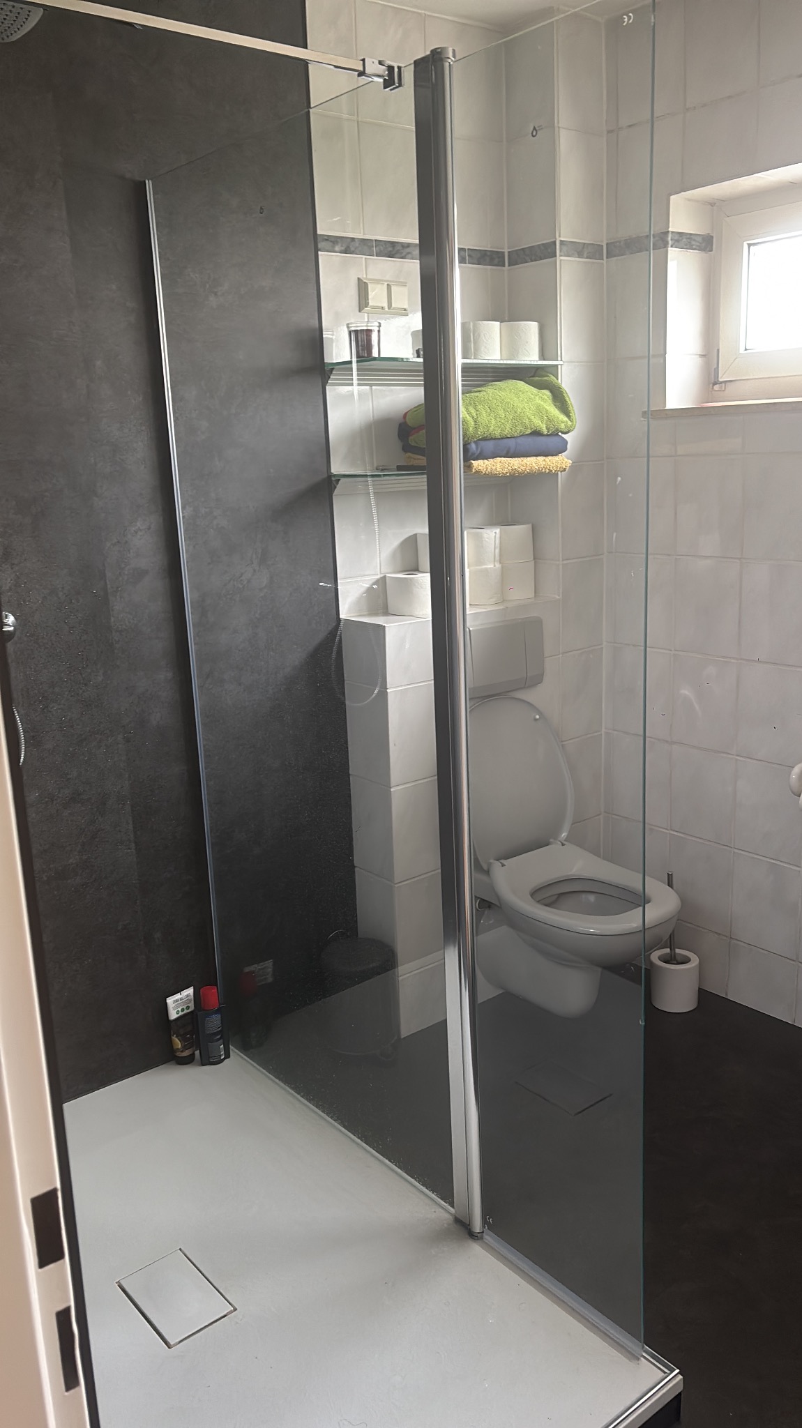 Nowoczesna kabina prysznicowa walk-in z grafitową ścianą i białymi płytkami. Widoczne szklane drzwi, toaleta i półka z ręcznikami. Minimalistyczny design łazienki.