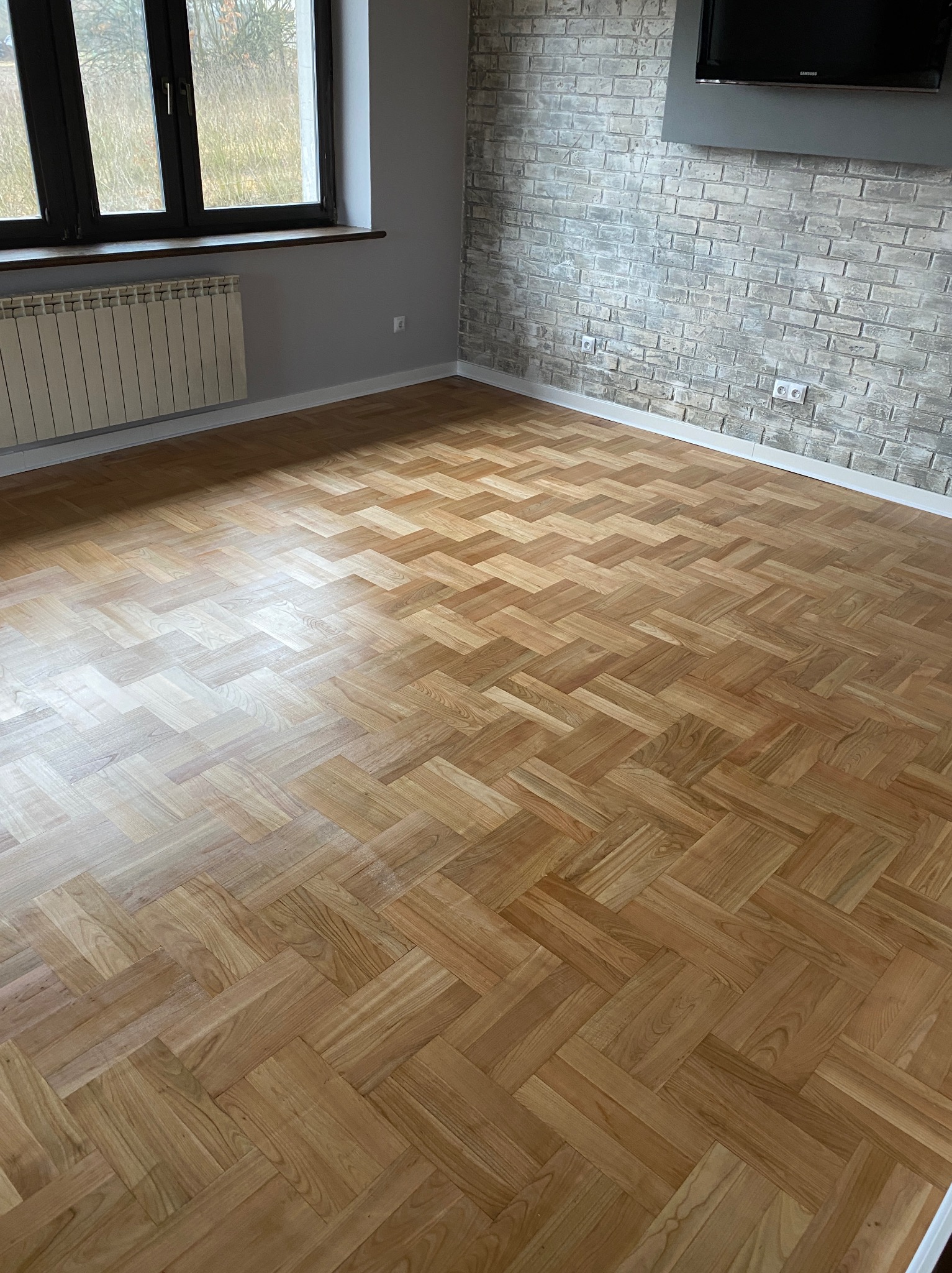 Parkiet ułożony w jodełkę w jasnym pokoju z oknem i ścianą z cegły. Widoczny grzejnik i telewizor na ścianie. Całość w neutralnej kolorystyce.