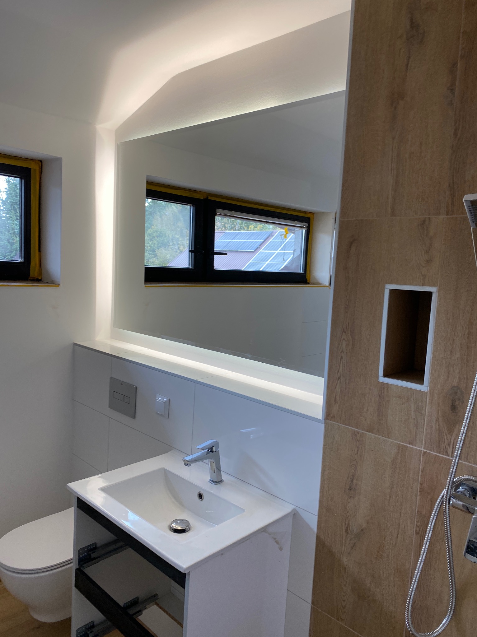 Nowoczesna łazienka z oświetlonym lustrem, umywalką na szafce, toaletą i kabiną prysznicową wyłożoną płytkami imitującymi drewno. Okno z widokiem na zewnątrz.