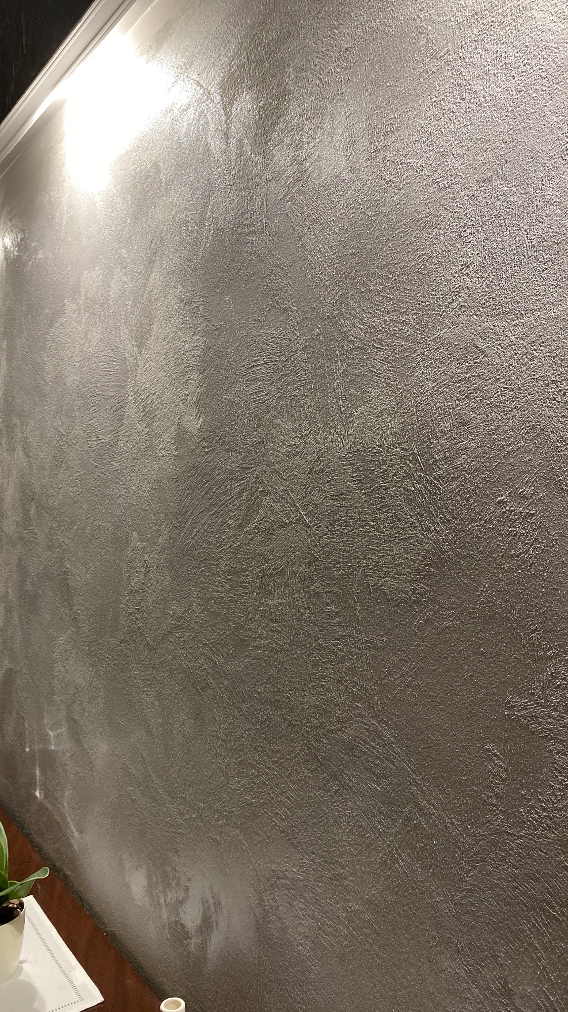Ściana z dekoracyjną, srebrzystą gładzią w pionowym ujęciu, oświetlona od góry, z widocznym fragmentem listwy przysufitowej i rośliną w doniczce w dolnym rogu.