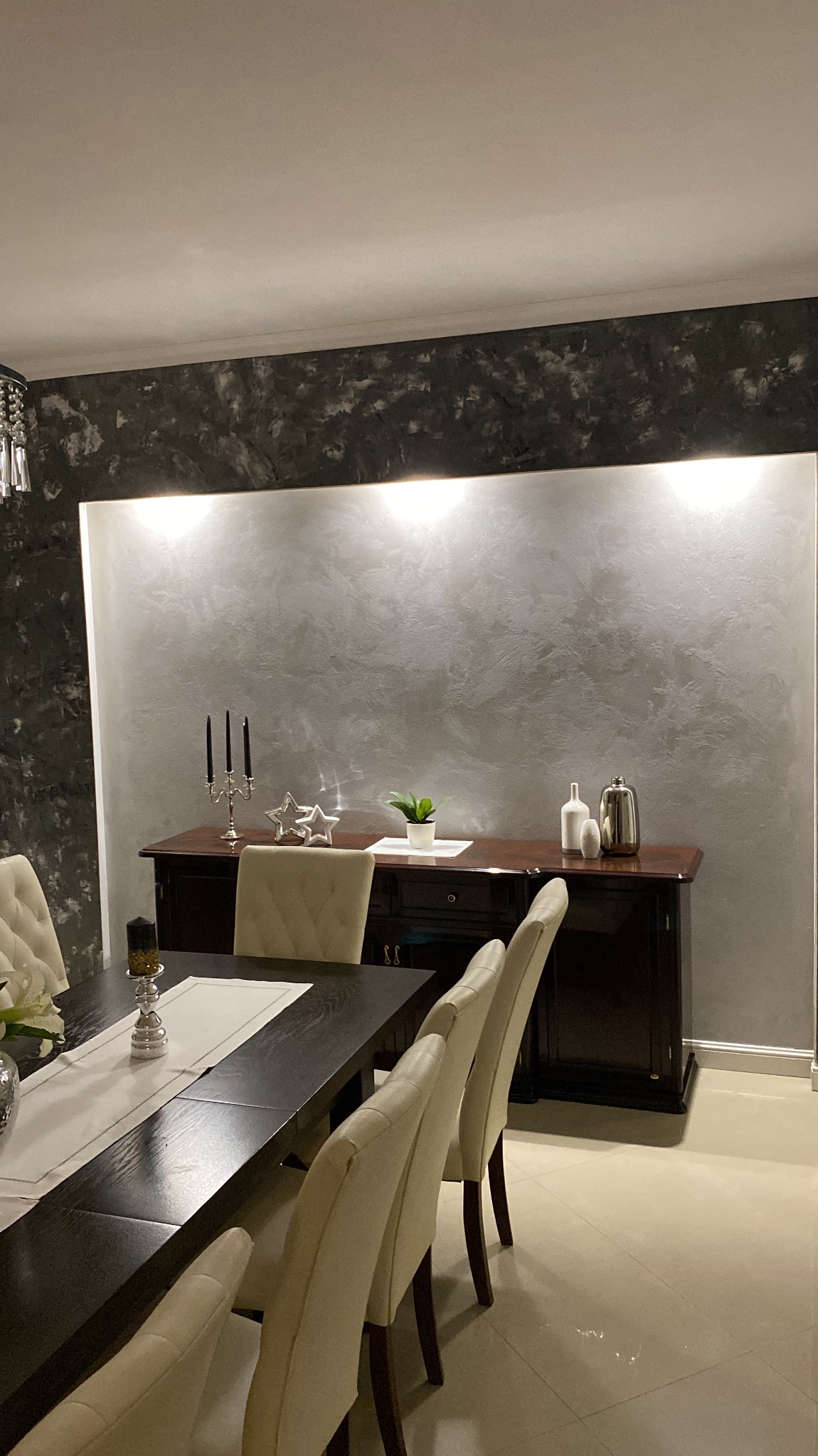 Eleganckie wnętrze jadalni z dekoracyjną ścianą w odcieniach szarości i czerni, oświetloną punktowo, z ciemnym kredensem i stołem z krzesłami w jasnej tonacji.