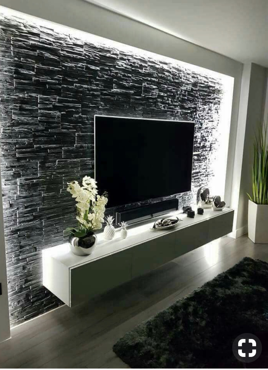 Ściana z czarnego kamienia dekoracyjnego z podświetleniem LED, na której wisi telewizor. Pod nim szafka RTV z dekoracjami. Nowoczesny design wnętrza.