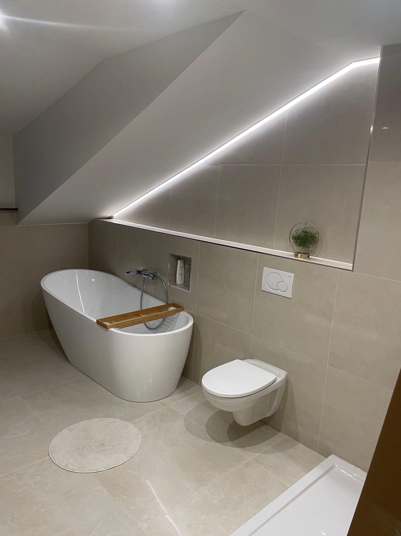 Nowoczesna łazienka na poddaszu z wanną wolnostojącą, podwieszaną toaletą i subtelnym oświetleniem LED. Beżowe płytki i minimalistyczny design.