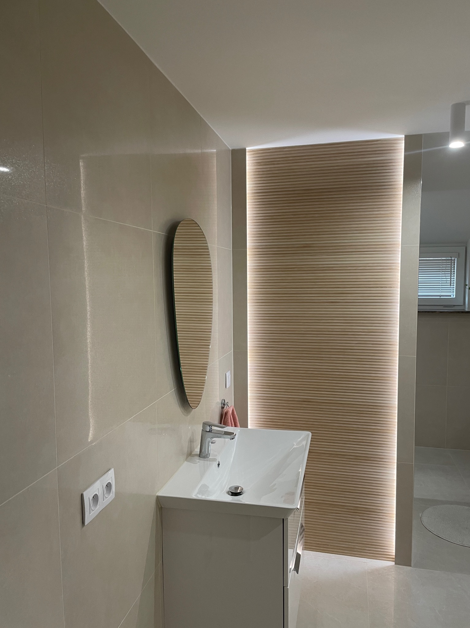 Jasna łazienka z owalnym lustrem, białą umywalką i drewnianą ścianką z podświetleniem LED. Minimalistyczny design, beżowe płytki, nowoczesne wykończenie.