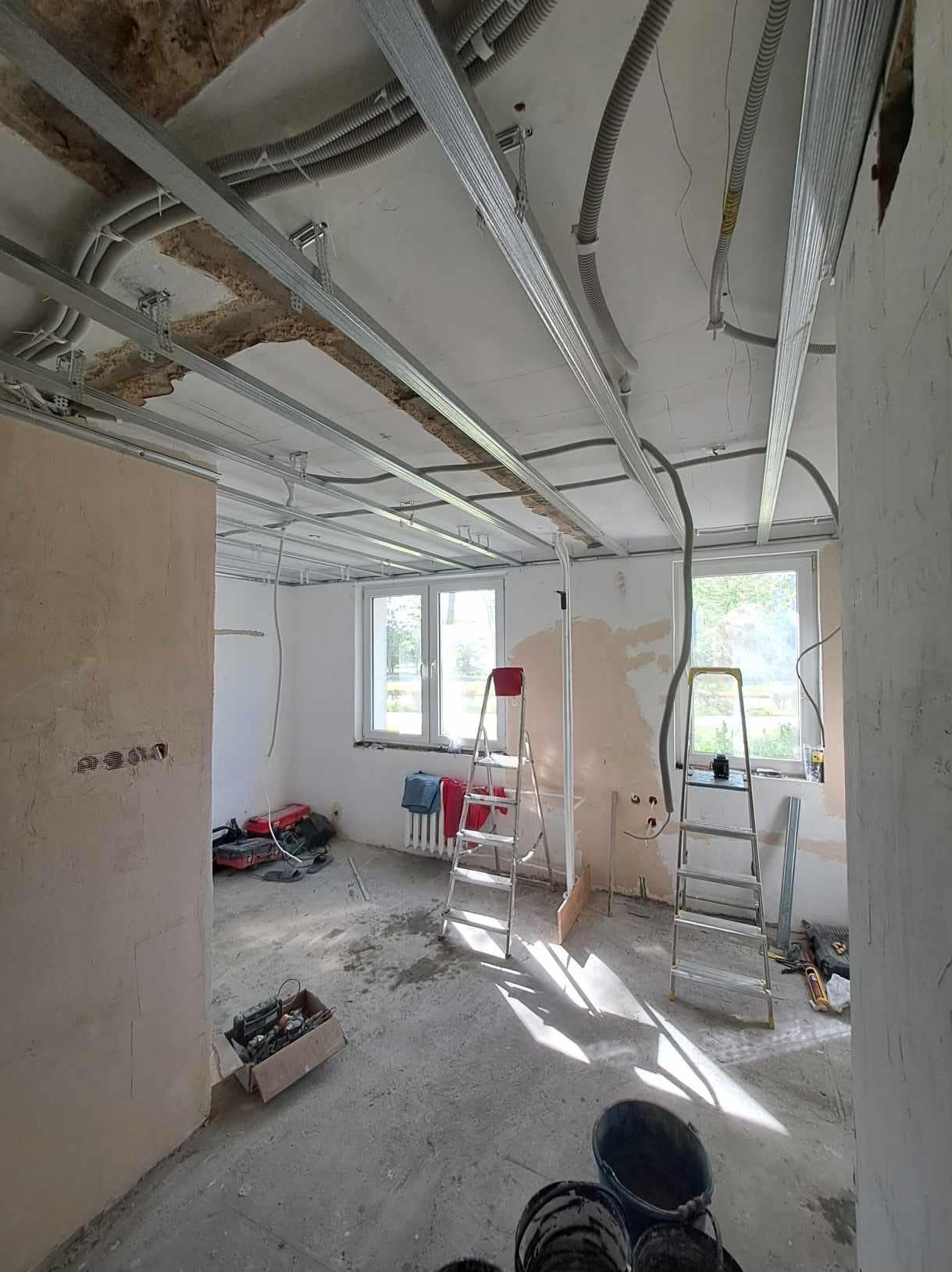 Wnętrze w trakcie remontu: widoczne profile sufitowe, instalacje elektryczne w peszlach, drabiny, narzędzia. Ściany przygotowane do malowania, okna z widokiem na zewnątrz.