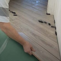 SVEET PRO - Układanie paneli podłogowych w jodełkę. Na podkładzie leżą panele, a przy ścianie umieszczone są czarne kliny dystansowe. Widoczne dłonie osoby montującej.