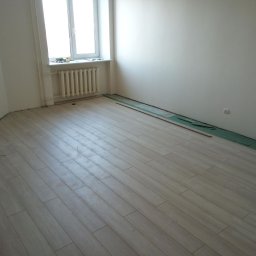 SVEET PRO - Jasne panele podłogowe ułożone w pokoju z oknem i grzejnikiem. Przy ścianach widoczne zielone podkładki i listwy przypodłogowe. Narzędzie do cięcia paneli leży na podłodze.