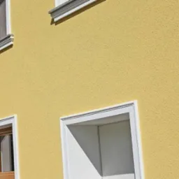 Elewacja budynku w odcieniach ż&oacute;łci z białymi obramowaniami okien i dekoracyjnym detalem przy dachu. Widoczny fragment zieleni w tle.