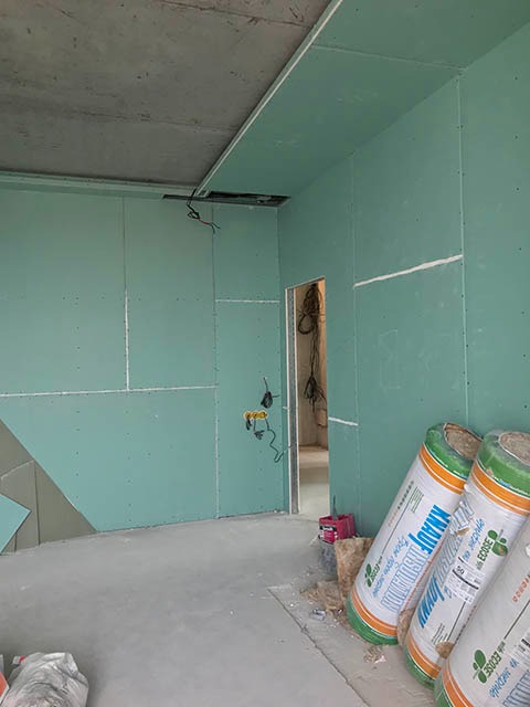 Wnętrze w trakcie wykańczania: ściany pokryte płytami gipsowo-kartonowymi, widoczne przewody elektryczne, narzędzia i materiały izolacyjne Knauf. Surowy betonowy sufit.