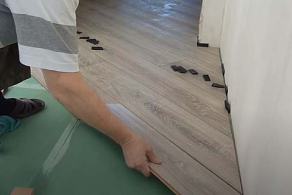 Układanie paneli podłogowych w jodełkę. Na podkładzie leżą panele, a przy ścianie umieszczone są czarne kliny dystansowe. Widoczne dłonie osoby montującej.