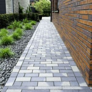 Długa ścieżka z szarej i białej kostki brukowej prowadząca wzdłuż ceglanego budynku, obok trawnika z krzewami i kamieniami ozdobnymi.