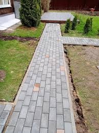 Ułożona ścieżka z szarej kostki brukowej w ogrodzie, z widocznym trawnikiem po bokach i fragmentem drewnianego tarasu w tle. Widoczne świeże nasadzenia roślin.