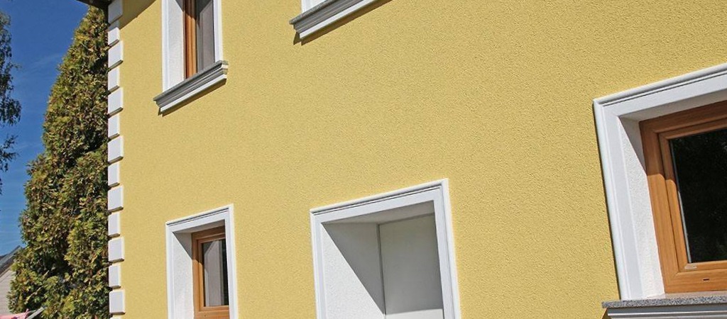 Elewacja budynku w odcieniach żółci z białymi obramowaniami okien i dekoracyjnym detalem przy dachu. Widoczny fragment zieleni w tle.