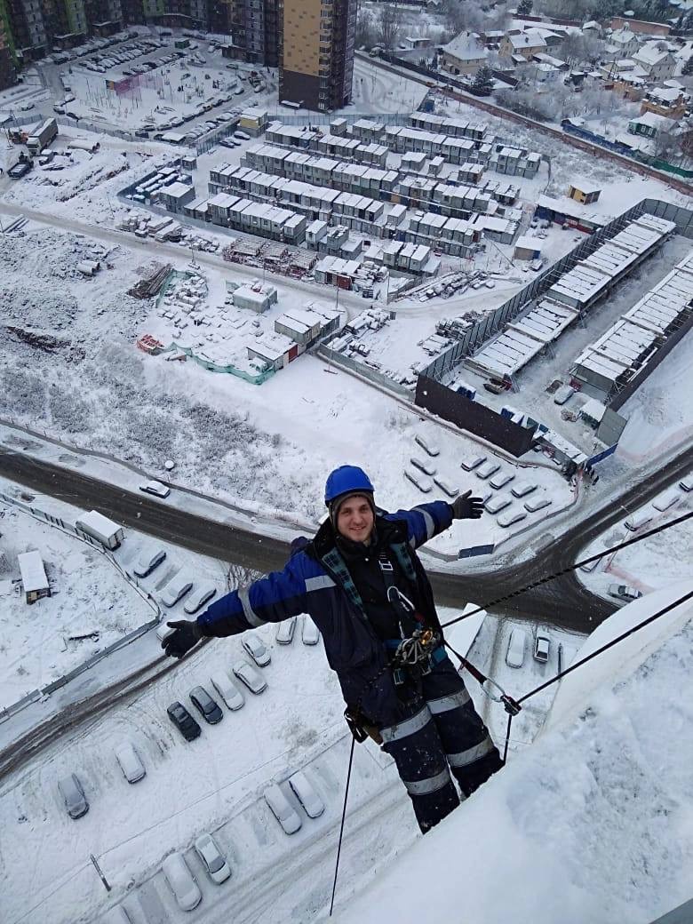 Pracownik w kasku, na linie, na tle zimowego krajobrazu z budynkami i parkingiem, uśmiechnięty, z rozłożonymi rękoma. Praca na wysokościach.