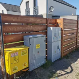Drewniane ogrodzenie posesji z żółtą skrzynką gazową i szarą szafką elektryczną na tle nowoczesnego domu z balkonami i anteną satelitarną.