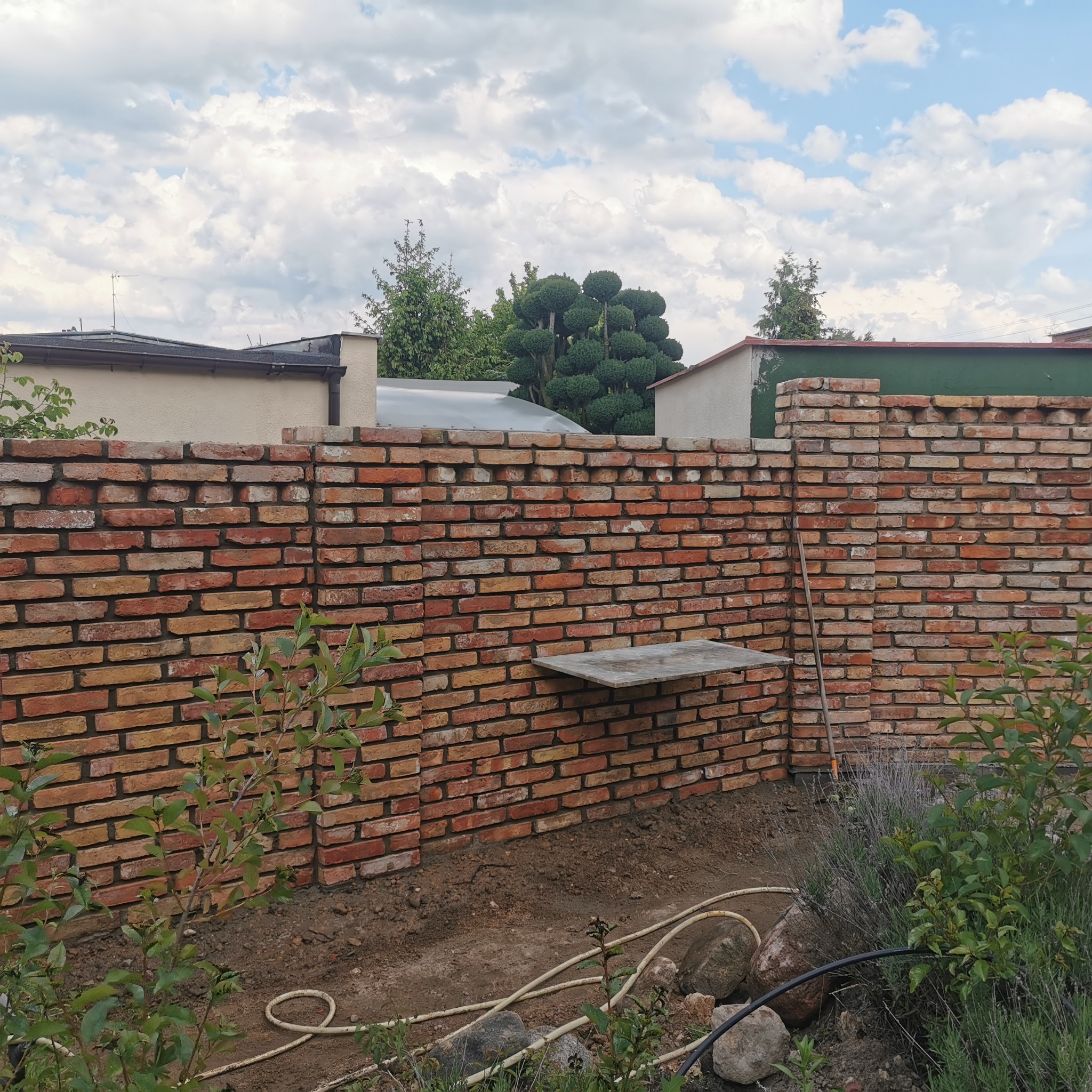 Ceglany mur ogrodzeniowy w trakcie budowy, z widoczną półką i elementami krajobrazu w tle, wykonany z czerwonej cegły, w słoneczny dzień.